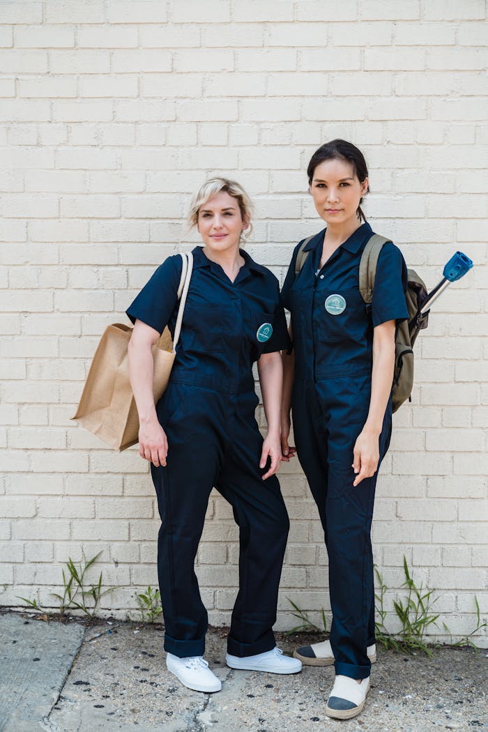 Two female workers in uniform with eco bags, standing against a white brick wall.